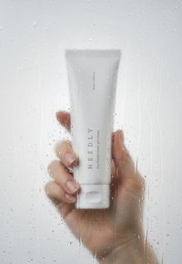 Tube blanc de crème gel à l'eau Panthénol NEEDLY de 50 ml, tenu à travers une surface givrée avec des gouttelettes, présentant un design simple et épuré avec du texte.