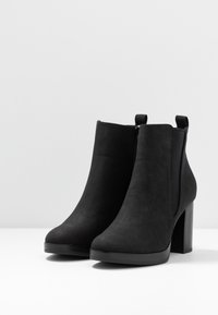 Botines de tacón alto negros de ante, con una textura suave, paneles elásticos laterales y un tacón cuadrado elevado.