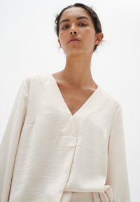 Helle Bluse mit V-Ausschnitt, langen Ärmeln und strukturiertem Finish. Verfügt über eine subtile Seitennähte-Tasche; der glatte Stoff fällt elegant.