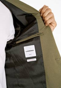 Schlanker Blazer in Olivgrün, mit einem schwarzen Satinfutter, Innenlabel-Details und einem eleganten, strukturierten Stofffinish.