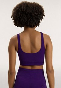 Mujer con cabello rizado que lleva un top corto sin mangas de color púrpura y leggings a juego de cintura alta, mostrada de espaldas sobre un fondo liso.