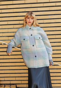 Veste checkered surdimensionnée bleu clair et crème avec deux poches avant, boutons noirs, portée sur une jupe sombre, devant un mur en briques décoré.