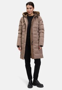 Beige polstret jakke med hætte, der har lynlås og knaplukning, sidelommer og et quiltet design. Bæres over en sort top og bukser.