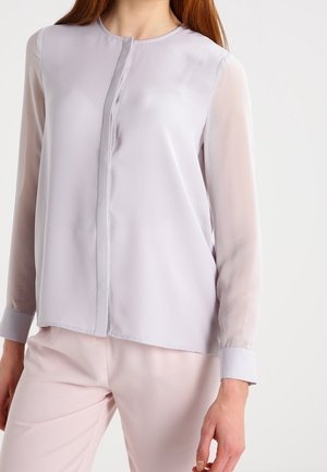 Nainen pukeutunut vaalean laventelinväriseen paitaan, jossa on piiloknapit ja läpikuultavat pitkät hihat, yhdistettynä vaaleanpunaisiin housuihin.