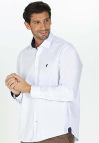 Chemise blanche à manches longues avec col bleu marine et petit logo noir sur la poitrine. Comprend une fermeture à boutons à l'avant et des poignets contrastés. Texture lisse.