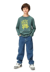 Grünes Langarm-T-Shirt mit gelbem Grafiktext; kombiniert mit blauen Jeans und weißen Sneakern mit schwarzen Akzenten.