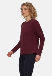 Kvinne med krøllete blondt hår som har på seg en maroon langermet skjorte og svarte leggings, stående sidelengs mot en ensfarget bakgrunn.