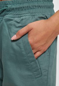 Tyrkysové, bavlněné joggers kalhoty s elastickým pasem a šňůrkou; mají boční kapsu a hladký textilní povrch.