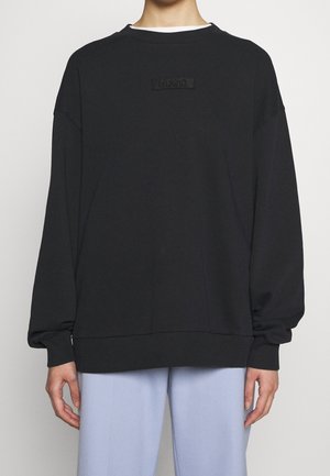 Personne portant un sweat-shirt noir oversize avec le logo "HUGO" centré sur la poitrine et un pantalon ample bleu clair.