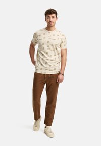Beige T-Shirt mit kurzen Ärmeln und strukturiertem Muster, kombiniert mit braunen textured Hosen und beigen Sneakers. Einfaches Design, lässiger Schnitt.