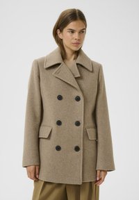 Cappotto in misto lana beige con design doppiopetto, ampie revers e cinque bottoni neri. Presenta due tasche frontali. Texture liscia.