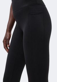 Gros plan sur des leggings noirs taille haute avec poche latérale portés par une personne à la peau foncée, montrant une main reposant près de la cuisse.