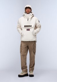 Beige waterdichte jas met voorzak en logo, gecombineerd met losse kaki cargobroeken en off-white wandelschoenen met groene veters.
