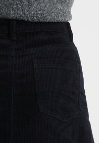 Gros plan sur un pantalon en velours côtelé noir avec une poche arrière, porté avec un pull en tricot gris rentré.