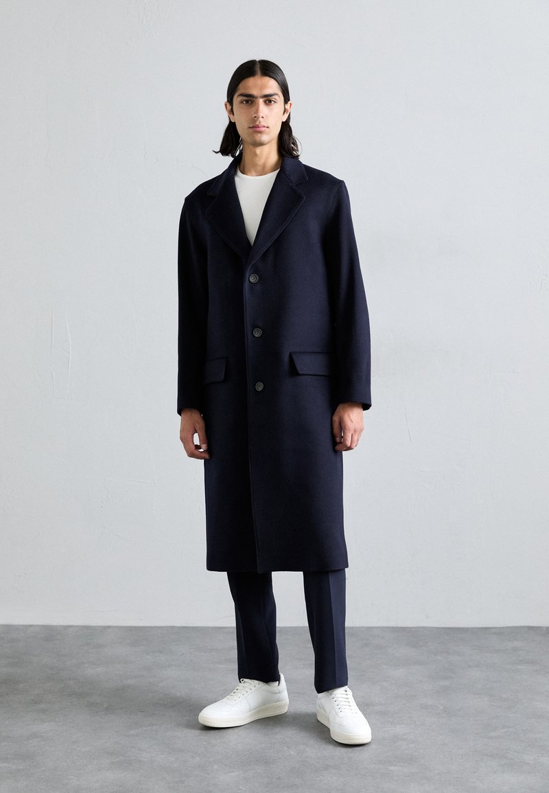 Cappotto di lana blu navy con revers a pacchetto, chiusura con bottoni e tasche frontali, abbinato a pantaloni scuri e sneakers bianche su un pavimento grigio.
