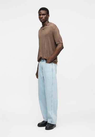 Homme debout les mains dans les poches, portant une chemise marron texturée à manches courtes, un jean large bleu clair et des chaussures noires sur fond blanc.