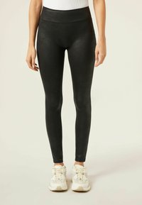 Schwarze Leggings mit glänzender Textur, hoher Taille und figurbetontem Design. Kombiniert mit weißen Sport Schuhen.