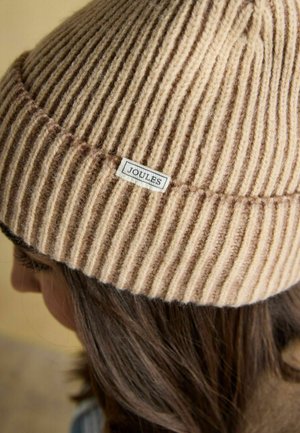 Beige gerippt gestrickte Mütze mit einem kleinen "Joules"-Etikett, getragen von einer Person mit braunen Haaren, von der Seite und leicht von oben betrachtet.