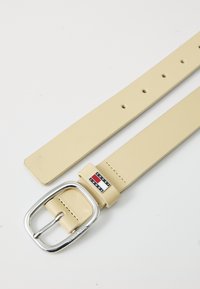 Beige syntetisk lærbelte med en sølvfarget metallspenne, som har et logomerke i rødt, hvitt og blått nær spennen.