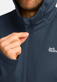 Giacca zip-up blu navy con colletto alto, caratterizzata da una superficie liscia, un logo discreto sul petto e un tirante sulla cerniera.