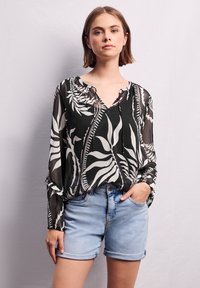 Blusa bianca e nera con motivi a foglia, tessuto trasparente, maniche lunghe con polsini arricciati, scollatura a V, abbinata a pantaloni corti in denim azzurro.