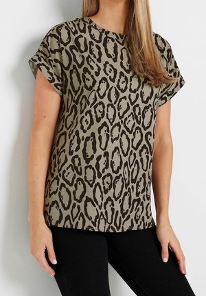 Femme portant un haut à manches courtes, ample, avec un imprimé animalier abstrait beige et noir, associé à un pantalon noir.
