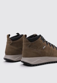 Botas de senderismo de ante marrón con una suela de goma gris, que presentan cordones negros y detalles laterales, diseñadas para la durabilidad y la comodidad.