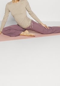 Beige long-sleeve top with side ruching, paired with loose, ankle-length mauve pants. Model poses on a pink mat, barefoot.
