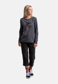 Camicia grigia a maniche lunghe con una grafica, abbinata a pantaloni neri cropped e sneakers nere. Calzini a strisce rosse e grigie visibili.