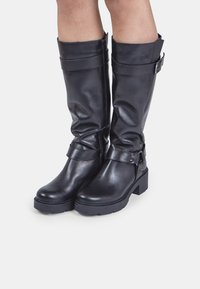 Botas de cuero negro hasta la rodilla con un diseño elegante, que cuentan con un tacón grueso, detalles de hebilla y un cierre de cremallera en la parte posterior.
