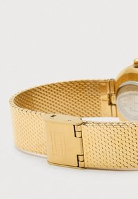 Χρυσό πλέγμα ρολογιού με λεία, ευέλικτη υφή. Διαθέτει κλείσιμο με κούμπωμα και κυκλικό καντράν ρολογιού, που είναι μερικώς ορατό.