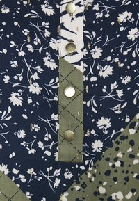 Blommönstrad tyg med sektioner i marinblått, vitt och olivgrönt. Guldiga detaljer på knapparna; broderade blommor och prickiga detaljer.