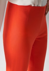 Pantalon rouge ajusté en tissu lisse, avec une taille haute aux lignes épurées et un minimum de détails. La couleur est vive et uniforme.