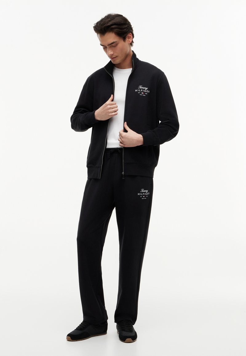 Mann trägt schwarzen Tommy Hilfiger Trainingsanzug mit weißem Logo, zieht den Reißverschluss der Jacke über einem weißen T-Shirt hoch, steht vor einfachem weißem Hintergrund.