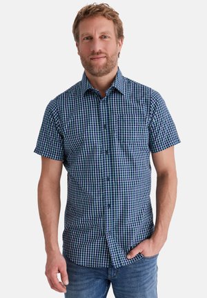 Mann trägt ein blau-grün kariertes, kurzärmeliges Hemd mit Knöpfen, eine Hand in der Jeanstasche, steht vor einem weißen Hintergrund.