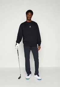 Μαύρη φούτερ με λογότυπο, σκούρα μπλε παντελόνια, λευκά παπούτσια γκολφ με μπλε λεπτομέρειες. Το μοντέλο κρατά ένα μπαστούνι γκολφ και φοράει ένα λευκό γάντι γκολφ.