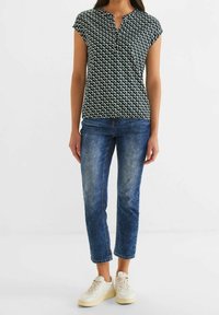 Krátký rukáv blůza tmavě zelené barvy s bílým a světle zeleným geometrickým vzorem, kombinovaná se světle modrými džíny a bílými teniskami.