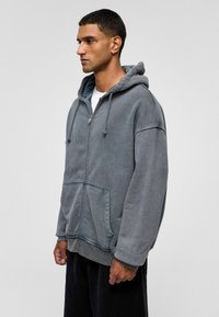 Grijze zip-up hoodie met een ruime capuchon, een voorzak en ribbels bij de mouwen. Gemaakt van een zachte stof met een licht versleten textuur.