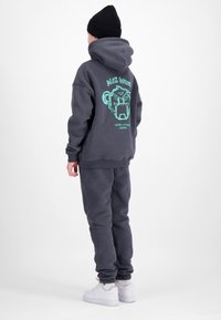 Persoon met een donkergrijze hoodie met een turkooizen afbeelding en tekst, bijpassende joggingbroek, witte sneakers en een zwarte gebreide muts, met de rug naar de camera op een effen achtergrond.