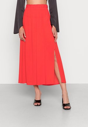 Red pleated maxi skirt with a side slit, elastic waistband, and smooth fabric. Paired with black open-toe heels, set against a plain background.