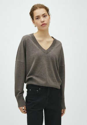 Jeune femme aux cheveux châtain clair attachés porte un pull ample marron à col en V et un pantalon noir sur un fond gris uni.