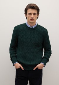 Maglione a treccia verde scuro con scollatura rotonda, polsi e orlo a coste, indossato sopra una camicia a righe blu chiaro.