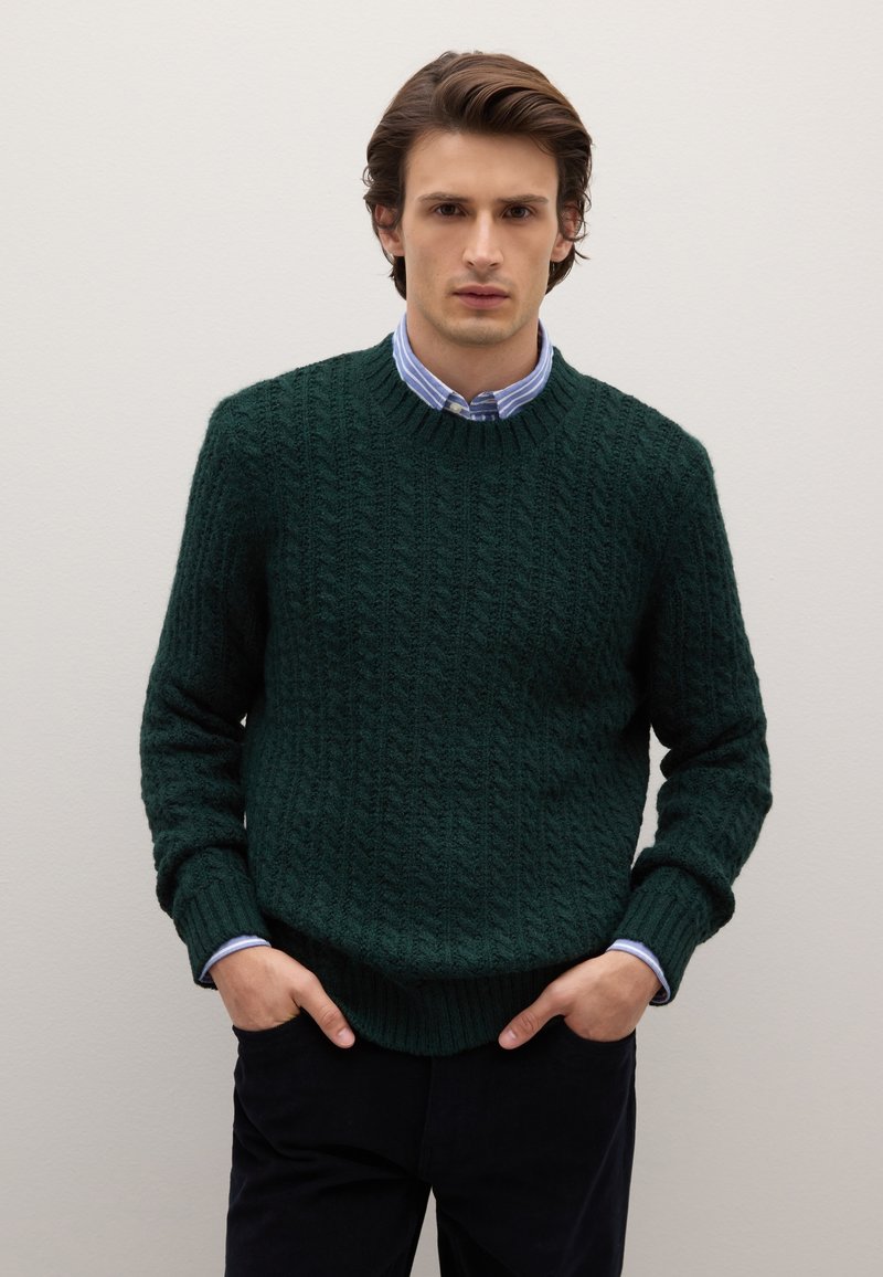 Maglione a treccia verde scuro con scollatura rotonda, polsi e orlo a coste, indossato sopra una camicia a righe blu chiaro.