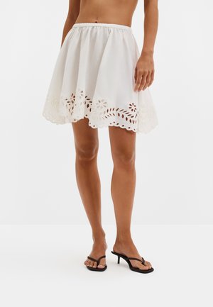 Femme portant une jupe blanche avec des découpes florales près de l'ourlet et des sandales noires à talons en forme de tongs, debout sur un fond blanc uni.