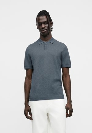 Schwarzer Mann mit geflochtenem Haar, der ein strukturiertes blaues Kurzarm-Poloshirt und weiße Hose trägt und vor einem einfarbigen hellen Hintergrund steht.