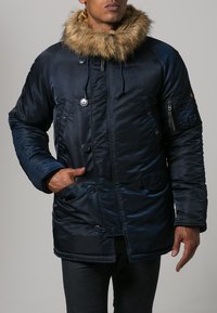 Cappotto isolato blu navy con cappuccio foderato in pelliccia, multiple bottoni, tasca con zip e texture trapuntata. Materiale in poliestere.