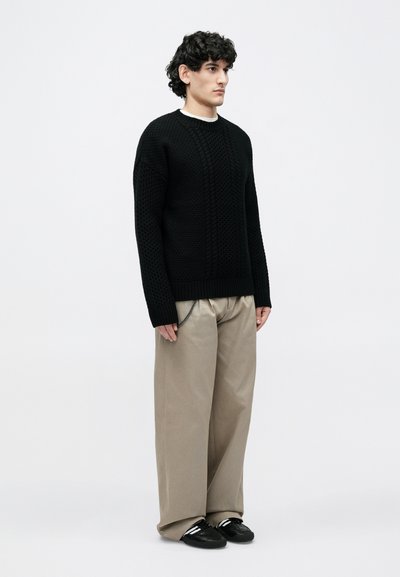 Νεαρό άτομο με σγουρά μαλλιά, φοράει μαύρο πουλόβερ με υφή, μπεζ παντελόνια φαρδιάς γραμμής και μαύρα αθλητικά παπούτσια, στέκεται μπροστά σε ένα απλό φόντο.