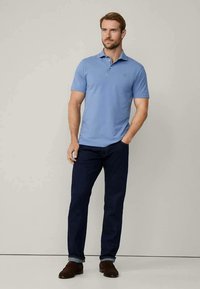 Vīrietis stāv pret vienkāršu fonu, ģērbies gaiši zilā īsajās piedurknēs polo krekla, tumši zilās džinsās un brūnās kurpēs, ar vienu roku kabatā.