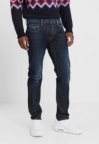 Mörkblå denimjeans med slim fit, fem fickor och synlig sömnad. Bärs med en mönstrad tröja och vita sneakers.