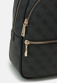 Esquina de una mochila negra texturizada con cremalleras doradas y un patrón en relieve, mostrando los compartimentos delantero y lateral.